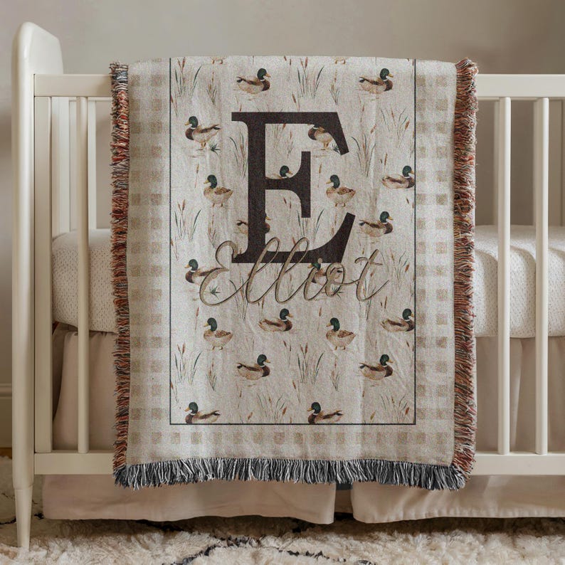 May include: A woven throw blanket featuring a repeating pattern of mallard ducks and reeds on a beige background. The center displays a large dark brown letter E with the name Elliot written in cursive script below it. The edges have a fringe.