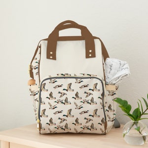 May include: A beige and brown diaper bag with a duck print. The bag has a large main compartment and a smaller pocket on the front. The bag also has a brown handle and adjustable straps.