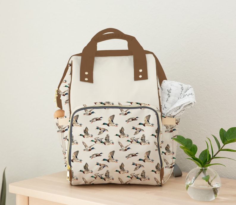 May include: A beige and brown diaper bag with a duck print. The bag has a large main compartment and a smaller pocket on the front. The bag also has a brown handle and adjustable straps.