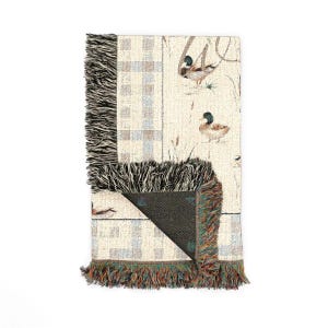 May include: A woven throw blanket featuring a cream-colored background with a repeating pattern of mallard ducks and tall grasses. The blanket has a plaid border design on one side and is finished with multi-colored fringed edges on the ends.