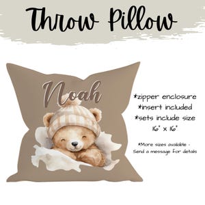May include: A taupe throw pillow with a watercolour illustration of a teddy bear wearing a striped hat. The pillow is personalised with the name "Noah". The pillow measures 40.6 cm x 40.6 cm.