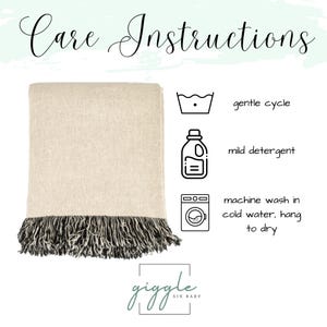 May include: A beige throw blanket with black fringe, featuring care instructions with icons for gentle cycle, mild detergent, and machine wash in cold water, hang to dry. The text Care Instructions and the brand name giggle SIX BABY are visible.