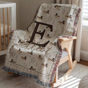 May include: A woven throw blanket featuring a repeating pattern of mallard ducks and reeds on a beige background. The center displays a large dark brown letter E with the name Elliot written in a cursive script font over the letter.