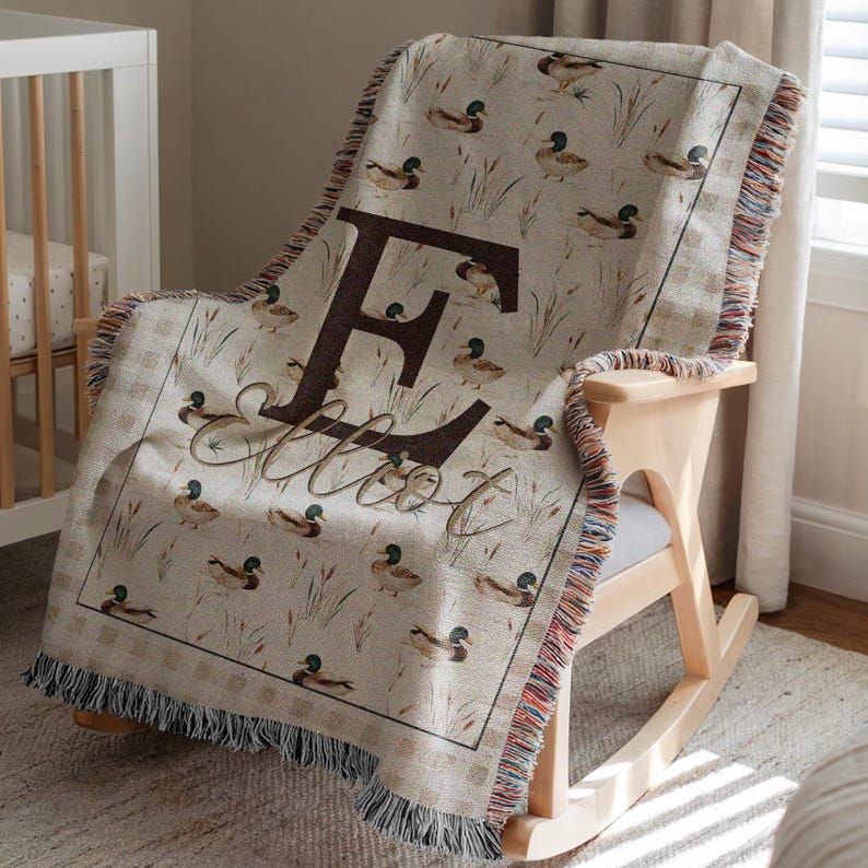 May include: A woven throw blanket featuring a repeating pattern of mallard ducks and reeds on a beige background. The center displays a large dark brown letter E with the name Elliot written in a cursive script font over the letter.