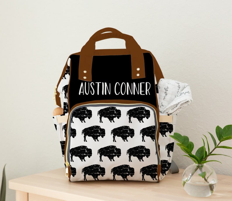 May include: A black and white diaper bag with a brown leather trim. The bag has a buffalo plaid pattern and the name "Austin Conner" is printed on the front.