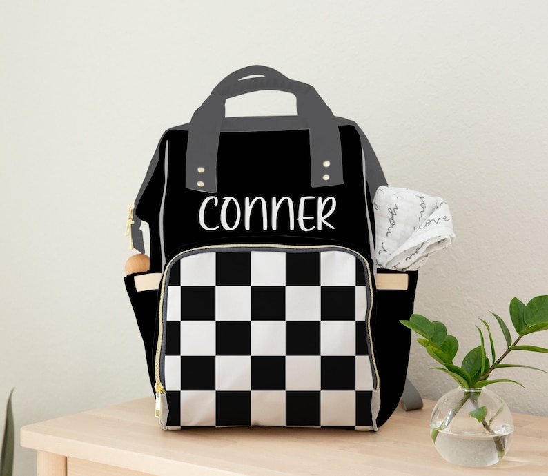 Puede incluir: Bolso de pa&ntilde;ales a cuadros blancos y negros con el nombre "CONNER" en escritura blanca. El bolso tiene un asa superior y ribetes grises, con cremalleras doradas y bolsillos laterales. Un pa&ntilde;o blanco est&aacute; metido en un bolsillo lateral.