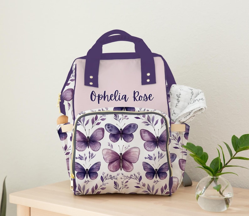 May include: Purple and white diaper bag backpack with a butterfly pattern and the name "Ophelia Rose" printed on the front.