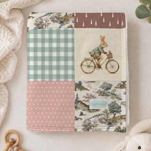 May include: A rectangular baby blanket featuring a patchwork design with six distinct panels. Patterns include a sage green gingham check, a dusty pink with white polka dots, a brown panel with white trees, and a landscape print with a rabbit on a bicycle.