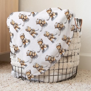 May include: A white blanket with a repeating pattern of brown teddy bears playing hockey. The blanket is folded and placed in a wire basket.