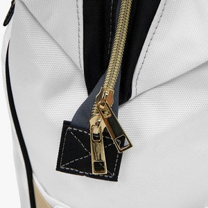 May include: Close-up of a white zipper with a gold zipper pull and black trim. The zipper is on a white fabric bag.