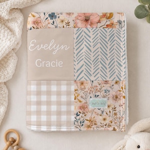 May include: A square baby blanket featuring a patchwork design with floral patterns, a beige and white gingham section, and a blue herringbone print. The beige panel is personalised with the white text Evelyn Gracie. A small brand label is attached.