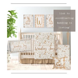 May include: A white crib with a brown and white floral patterned blanket and pillow. The pillow has the name "Wrenley Joy" embroidered on it. There are three framed prints above the crib, each featuring a bunny rabbit and floral designs. The prints have a white background and gold accents. The text "link to coordinating bedding in listing!" is in the upper right corner of the image.