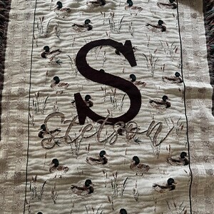 May include: A woven throw blanket featuring a repeating pattern of ducks and reeds on a beige background. The center displays a large dark brown letter S above the name Stetson in a cursive script. The blanket has fringed edges on the top and bottom.