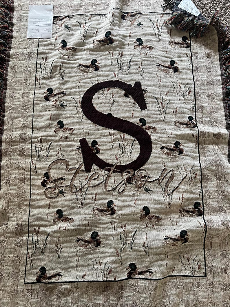 May include: A woven throw blanket featuring a repeating pattern of ducks and reeds on a beige background. The center displays a large dark brown letter S above the name Stetson in a cursive script. The blanket has fringed edges on the top and bottom.