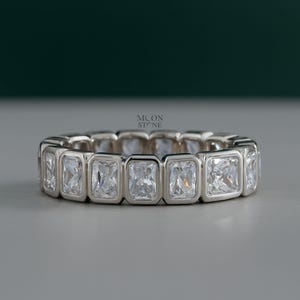 May include: A silver ring with rectangular, clear gemstones set in a bezel style. The ring features a continuous band of stones, creating a sparkling, elegant look. The brand name "MOON STONE" is visible.