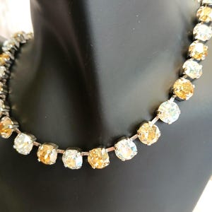 May include: A silver-toned necklace featuring alternating clear and amber-colored round crystals. The crystals are set closely together, creating a sparkling, elegant look. The necklace is displayed on a black mannequin.