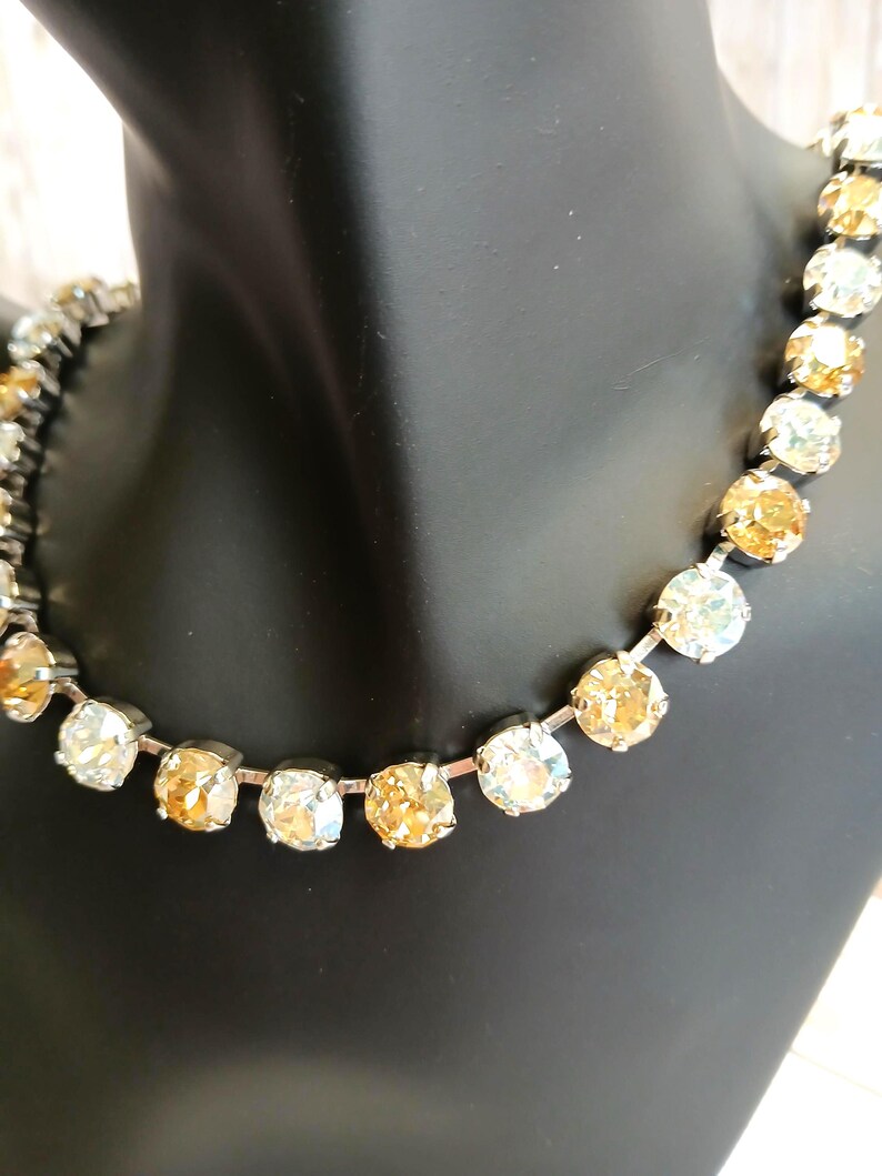 May include: A silver-toned necklace featuring alternating clear and amber-colored round crystals. The crystals are set closely together, creating a sparkling, elegant look. The necklace is displayed on a black mannequin.