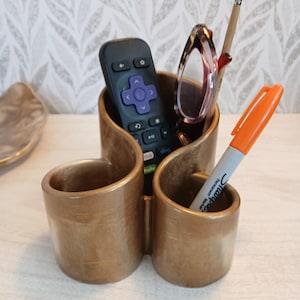 Puede incluir: Un organizador de escritorio de color dorado metálico con dos compartimentos y un diseño curvo en forma de S. Contiene un mando a distancia negro con un pad direccional morado, unas gafas de carey, un pincel fino y un rotulador Sharpie naranja.