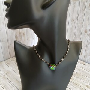 Peut inclure: Un collier &agrave; cha&icirc;ne argent&eacute; avec un pendentif rond et iris&eacute;. Le pendentif pr&eacute;sente un m&eacute;lange de teintes vertes, bleues et violettes. Le collier est pr&eacute;sent&eacute; sur un buste de mannequin noir, sur un fond en bois clair.