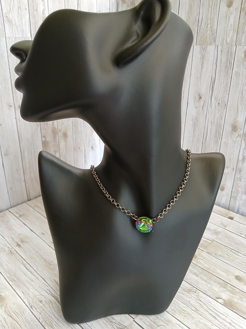 Peut inclure: Un collier &agrave; cha&icirc;ne argent&eacute; avec un pendentif rond et iris&eacute;. Le pendentif pr&eacute;sente un m&eacute;lange de teintes vertes, bleues et violettes. Le collier est pr&eacute;sent&eacute; sur un buste de mannequin noir, sur un fond en bois clair.