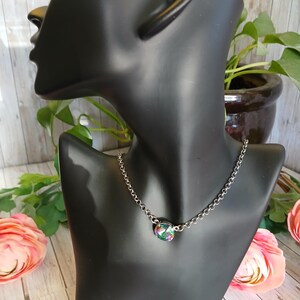 Peut inclure: Un collier &agrave; cha&icirc;ne argent&eacute;e avec un pendentif rond et iris&eacute;. Le pendentif affiche un m&eacute;lange de couleurs, dont le vert, le violet et le bleu. Le collier est pr&eacute;sent&eacute; sur un buste de mannequin noir, avec des fleurs et de la verdure en arri&egrave;re-plan.