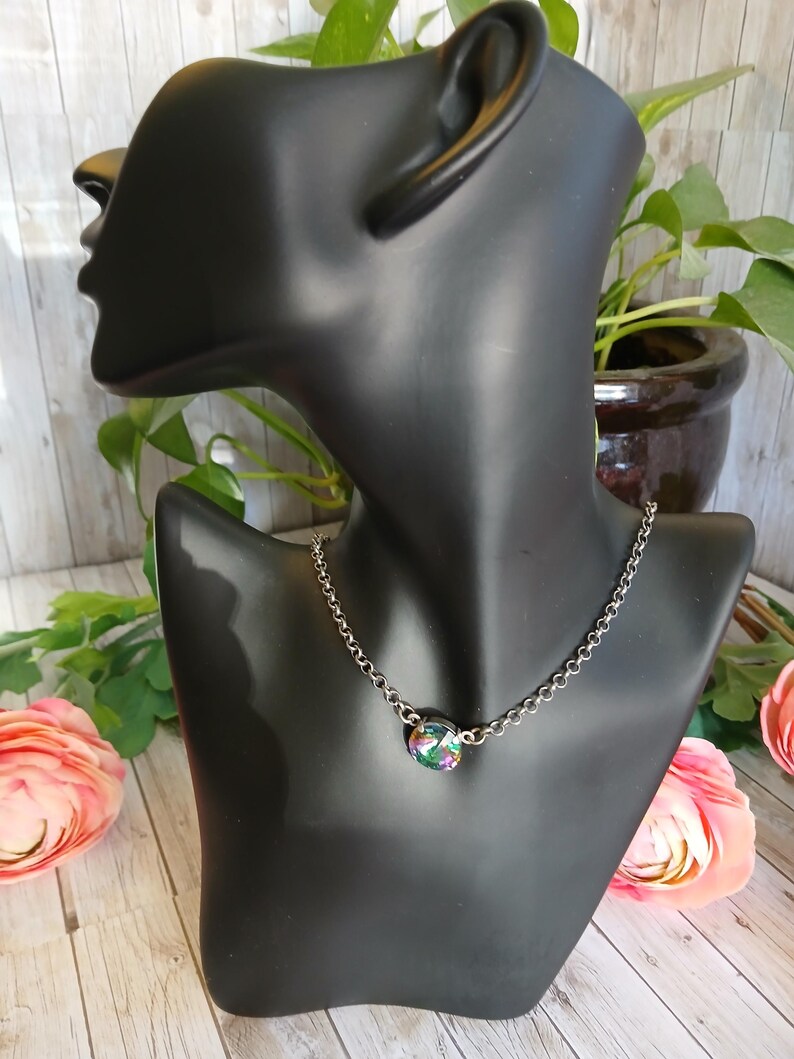 Peut inclure: Un collier &agrave; cha&icirc;ne argent&eacute;e avec un pendentif rond et iris&eacute;. Le pendentif affiche un m&eacute;lange de couleurs, dont le vert, le violet et le bleu. Le collier est pr&eacute;sent&eacute; sur un buste de mannequin noir, avec des fleurs et de la verdure en arri&egrave;re-plan.