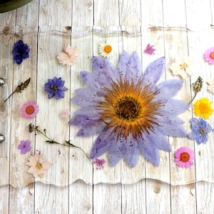 Pode incluir: Bandeja retangular transparente com borda ondulada, com flores secas em tons de roxo, rosa, amarelo e branco. A bandeja tem alças prateadas e está sobre um fundo de madeira clara.