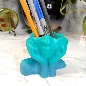 May include: A teal and blue pen holder shaped like a pair of hands cupped together to form a basin. The holder contains four markers, including orange, grey, blue, and black Sharpie permanent markers, standing upright within the central opening.