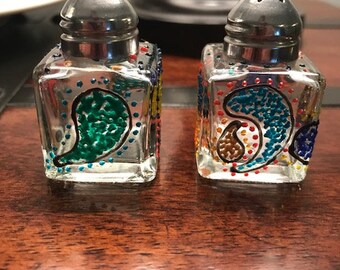 May include: Two small glass salt and pepper shakers with silver tops. The shakers are decorated with colorful paisley patterns and dots.