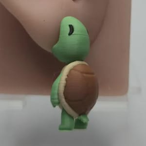 以下が含まれることがあります： 緑と茶色のカメのイヤリング。カメは緑色の頭と脚、茶色の甲羅、クリーム色のトリムをしています。イヤリングは肌色の耳のモデルに表示されています。