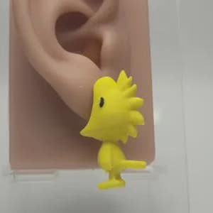 Könnte beinhalten: Ein gelber Cartoon-Vogel-Ohrring mit einem schwarzen Auge wird neben einem fleischfarbenen Ohr gezeigt. Der Vogel hat einen stacheligen Kopf und einen kleinen Schwanz. Der Ohrring ist ein lustiges, skurriles Accessoire.