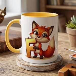 Può includere: Tazza in ceramica bianca con interno e manico gialli. La tazza presenta un personaggio di volpe dei cartoni animati che tiene la lettera "F". Sono visibili anche un sottobicchiere in legno e una piccola "F" in legno. Una statuina di volpe dorata e una pianta grassa sono sullo sfondo.