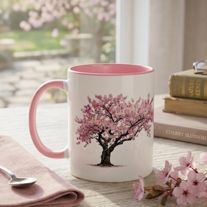 May include: A white ceramic mug with a pink interior and handle, featuring a detailed illustration of a blooming cherry blossom tree on the front. The mug is positioned next to a small silver spoon, a folded pink napkin, and a stack of books.
