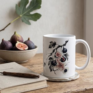 Puede incluir: Una taza de cerámica blanca con una ilustración botánica detallada de higos de color púrpura oscuro, fruta cortada y hojas verdes sobre una rama. La taza tiene un acabado liso y un asa curva, mostrando una representación realista de la fruta.