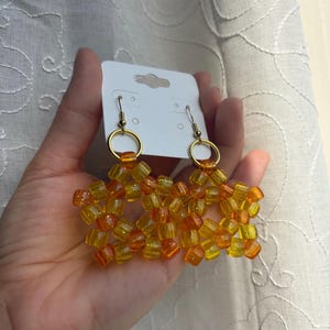 May include: A pair of star-shaped earrings crafted from translucent yellow and orange plastic pony beads. Each earring features a gold-toned metal jump ring connected to a fishhook ear wire, all displayed on a white rectangular card.