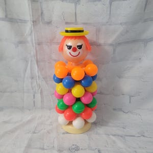 May include: A vintage clown toy with a yellow hat and orange hair. The clown is made of plastic and has a stack of colourful balls around its body.