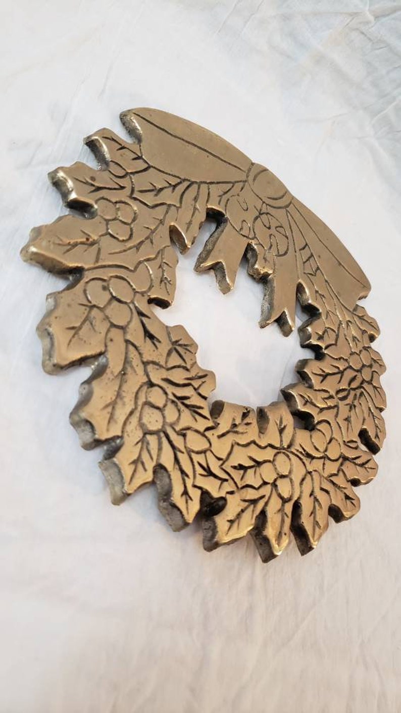 Pode incluir: Uma coroa detalhada, em tom bronze, com um design de folhas e bagas. A coroa &eacute; circular com um la&ccedil;o no topo. O metal tem uma apar&ecirc;ncia texturizada e envelhecida, sugerindo que &eacute; vintage ou antigo. A coroa &eacute; provavelmente usada para fins decorativos.