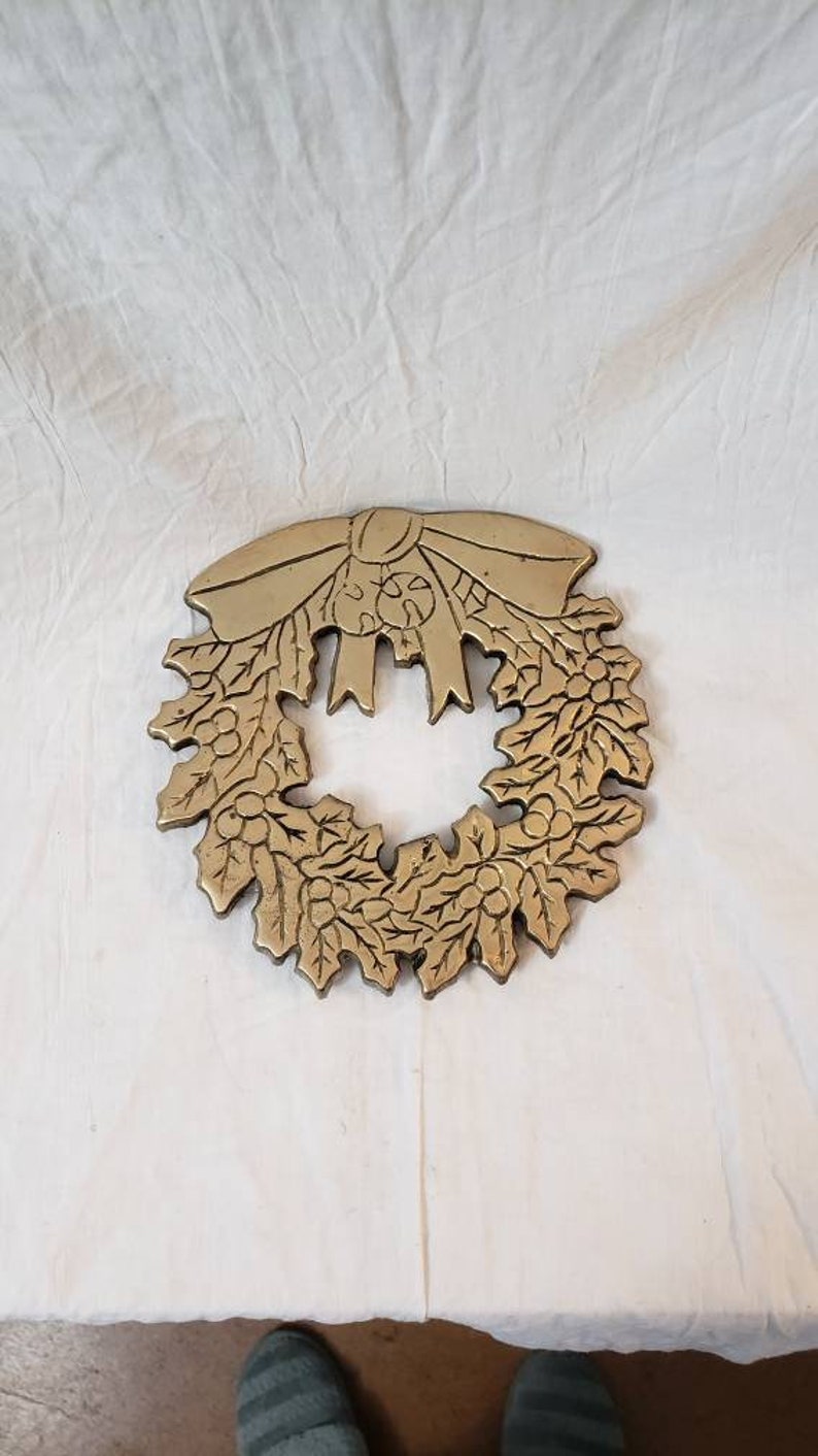 Pode incluir: Uma grinalda de Natal de metal dourado com um la&ccedil;o no topo. A grinalda apresenta folhas e bagas detalhadas, criando uma decora&ccedil;&atilde;o festiva. A grinalda tem uma forma circular.