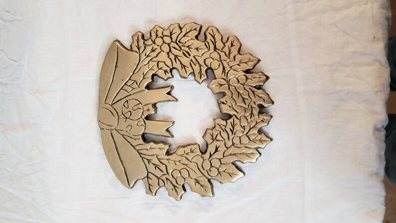 Pode incluir: Uma grinalda de metal dourado com um la&ccedil;o no lado esquerdo. A grinalda &eacute; decorada com folhas e bagas, e &eacute; provavelmente uma decora&ccedil;&atilde;o de Natal. A grinalda est&aacute; sobre uma superf&iacute;cie branca.