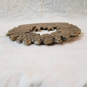 Pode incluir: Um objeto decorativo circular de cor bronze com uma superf&iacute;cie texturizada e detalhada. O objeto tem um design floral ou em forma de folha ao redor da borda externa e uma abertura central. &Eacute; exibido em um fundo branco texturizado.