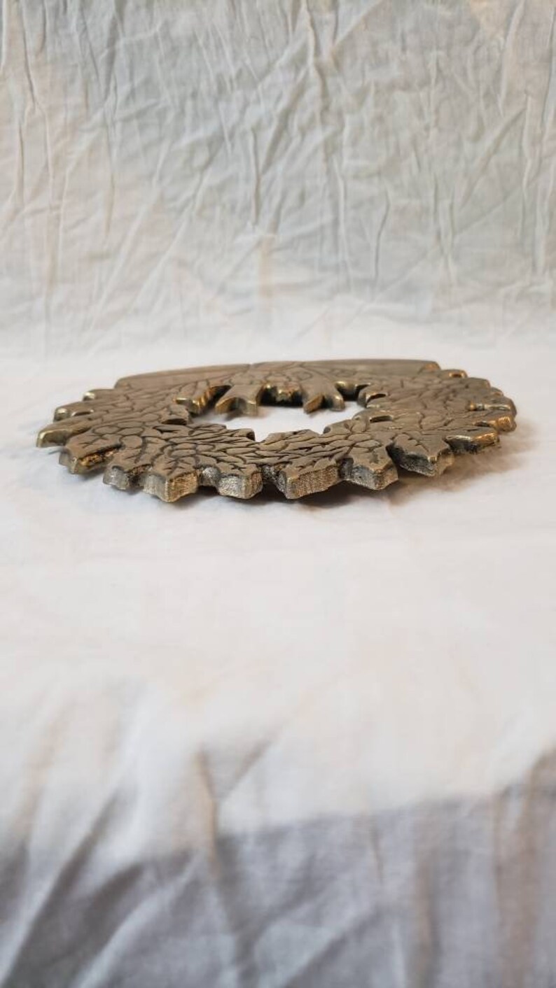 Pode incluir: Um objeto decorativo circular de cor bronze com uma superf&iacute;cie texturizada e detalhada. O objeto tem um design floral ou em forma de folha ao redor da borda externa e uma abertura central. &Eacute; exibido em um fundo branco texturizado.
