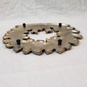 Pode incluir: Um descanso de panela de metal prateado com uma borda decorativa e irregular. O descanso de panela tem uma abertura central e quatro p&eacute;s de borracha pretos. O design lembra uma flor estilizada ou um raio de sol.