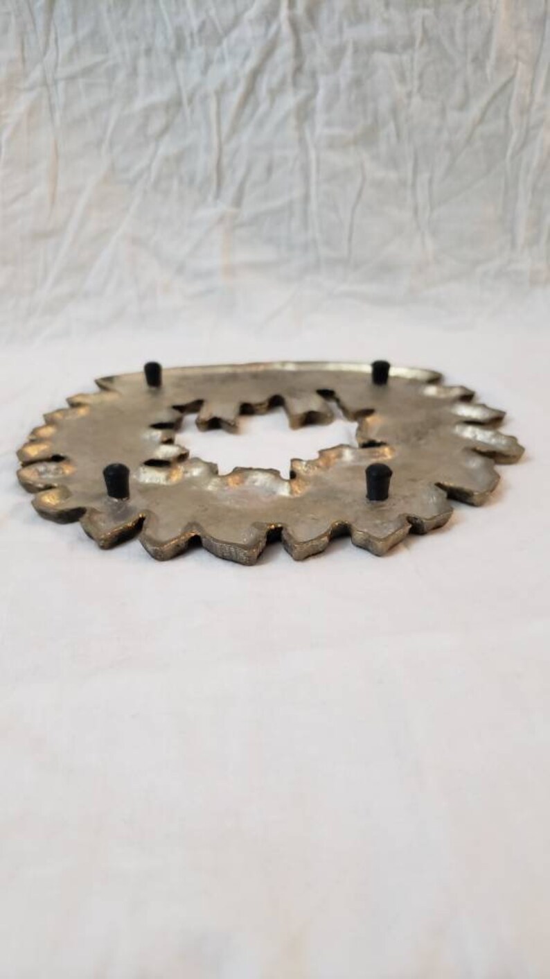 Pode incluir: Um descanso de panela de metal prateado com uma borda decorativa e irregular. O descanso de panela tem uma abertura central e quatro p&eacute;s de borracha pretos. O design lembra uma flor estilizada ou um raio de sol.