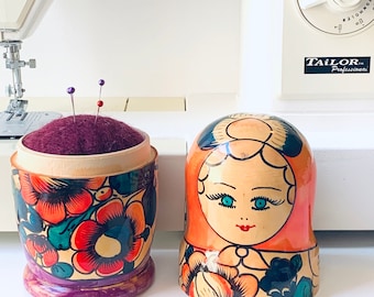Puede incluir: Dos muñecas rusas de madera, también conocidas como muñecas de anidación, con coloridos patrones florales. La muñeca más pequeña es un alfiletero con una parte superior de fieltro morado y está llena de alfileres. La muñeca más grande tiene una cara pintada con ojos azules y una boca roja. Las muñecas están sentadas sobre una superficie blanca.