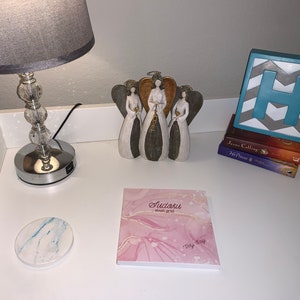 May include: A decorative arrangement on a white surface. It includes a silver-based lamp with a gray shade, a trio of angel figurines, a stack of books, a block with the letter "H", a marble coaster, and a pink Sudoku notepad.