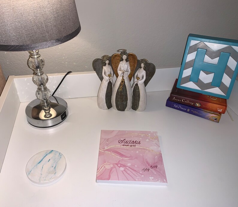 May include: A decorative arrangement on a white surface. It includes a silver-based lamp with a gray shade, a trio of angel figurines, a stack of books, a block with the letter "H", a marble coaster, and a pink Sudoku notepad.