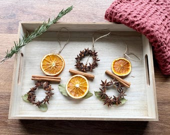 May include: A collection of handmade ornaments featuring dried orange slices, star anise wreaths, and cinnamon sticks. The ornaments are arranged on a light wooden tray alongside a sprig of fresh rosemary and a textured dark pink fabric.