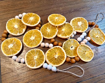 Puede incluir: Una guirnalda decorativa con rodajas de naranja secas intercaladas con cuentas de madera redondas en tonos blanco y marrón natural. Las rodajas están dispuestas a lo largo de un cordel de yute que forma bucles en ambos extremos.