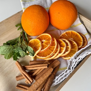 May include: A wooden tray displays two whole oranges, dried orange slices, fresh mint sprigs, and cinnamon sticks. A white and yellow striped cloth is under the oranges and slices. The oranges are bright orange, and the cinnamon sticks are brown.