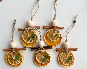 May include: Six handmade ornaments featuring dried orange slices, rosemary sprigs, and cinnamon sticks. Each ornament has a wooden bead and a twine loop for hanging. The orange slices are a vibrant orange color.